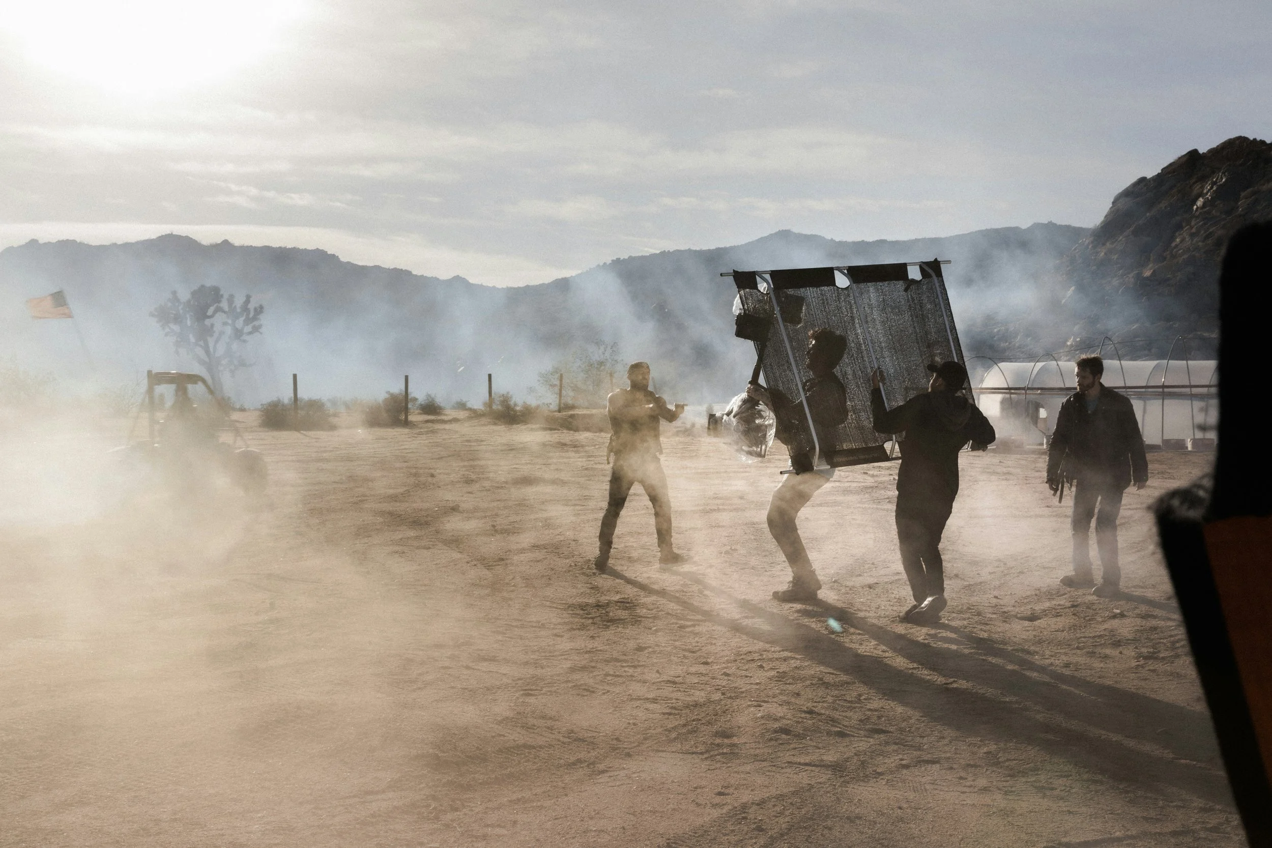 Film crew members working on set in dusty desert with mountains in background, some holding equipment, amidst smoke and haze.