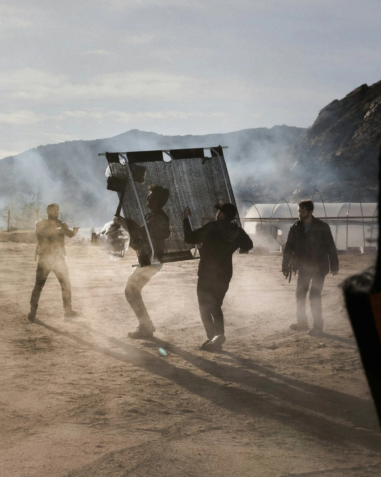 People working on a film set outdoors, carrying equipment and surrounded by dust and smoke, with mountains and greenhouses in the background.