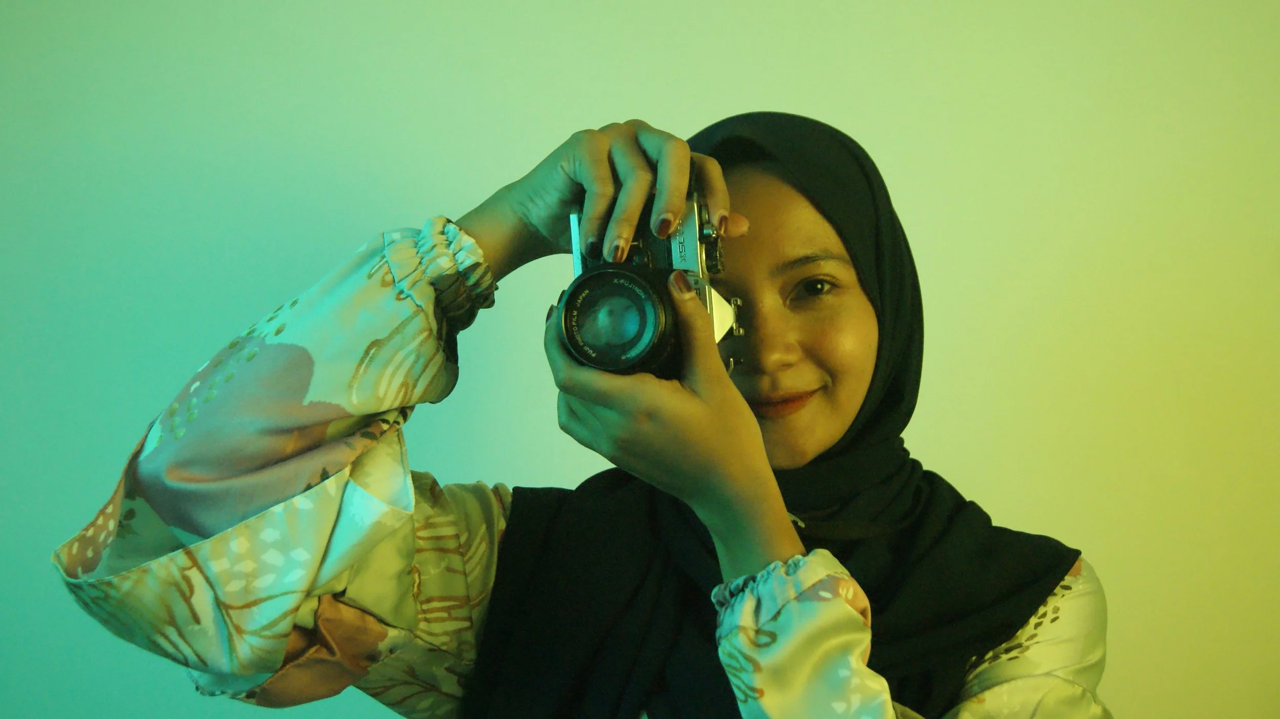 A young woman in a hijab taking a photo of herself with a vintage camera, smiling, against a gradient green background.