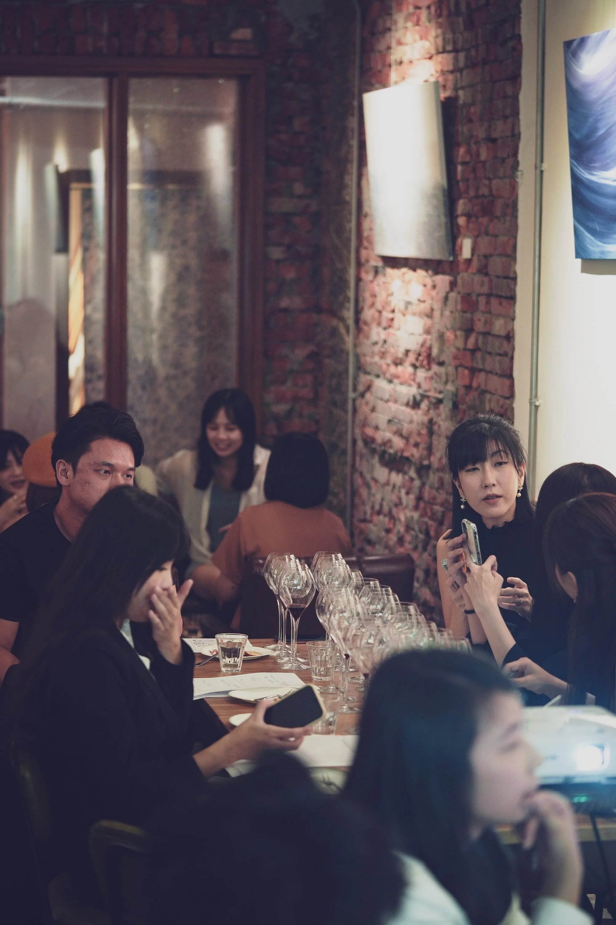 A group of people sitting around a dining table in a cozy restaurant with exposed brick walls, engaged in conversation and looking at their phones, and a woman gesturing as she speaks.