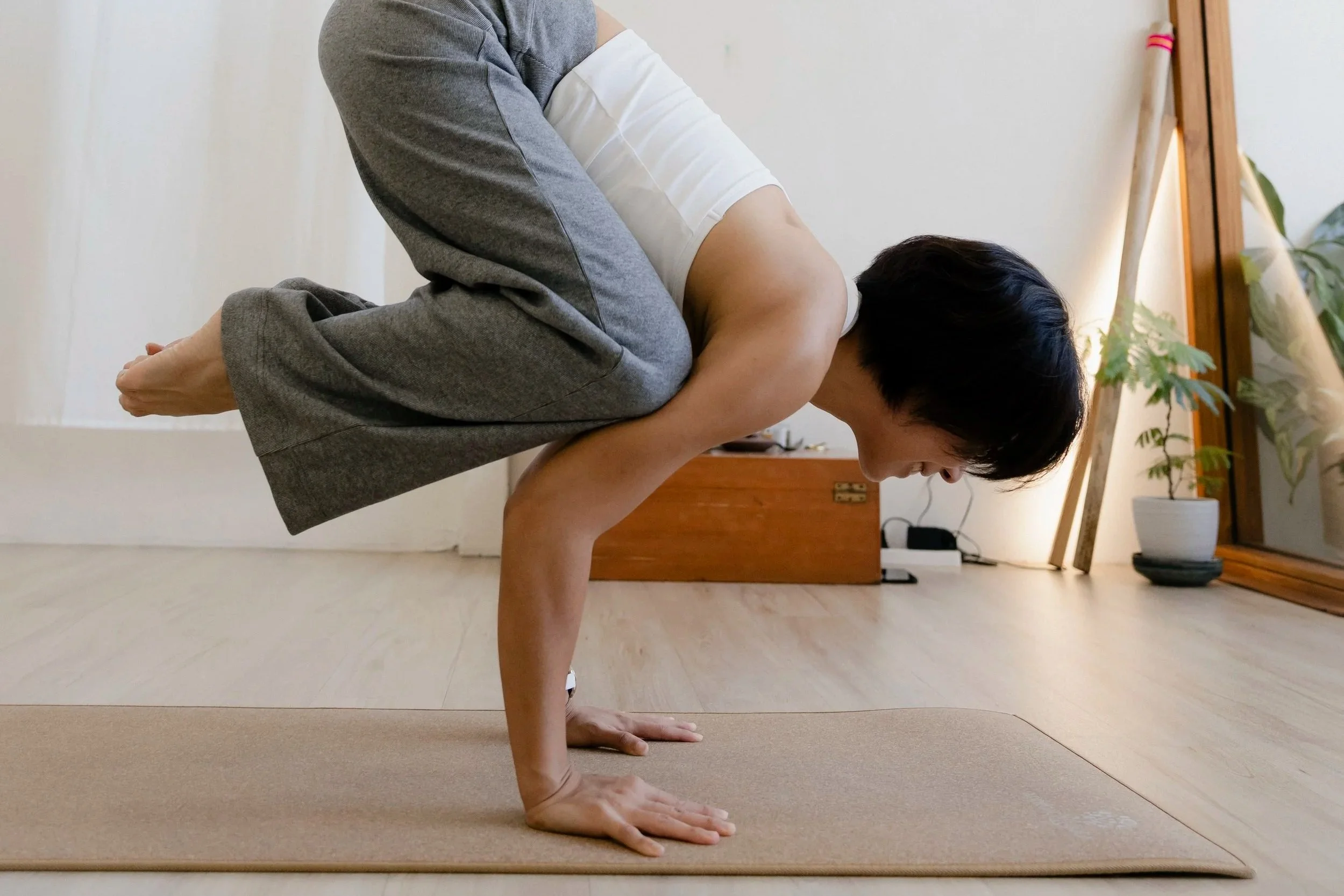 A woman with short black hair smiling, wearing a red spaghetti strap top, sitting on a wooden chair against a plain light-colored wall.