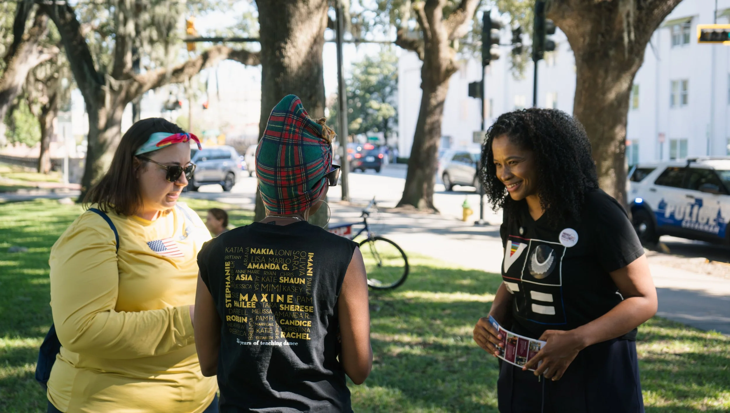 Three women standing in a park, engaged in conversation. The woman on the right is smiling and holding flyers, the woman in the middle is facing away with a colorful head wrap and glasses, and the woman on the left is wearing a yellow shirt, sunglasses, and a headband.