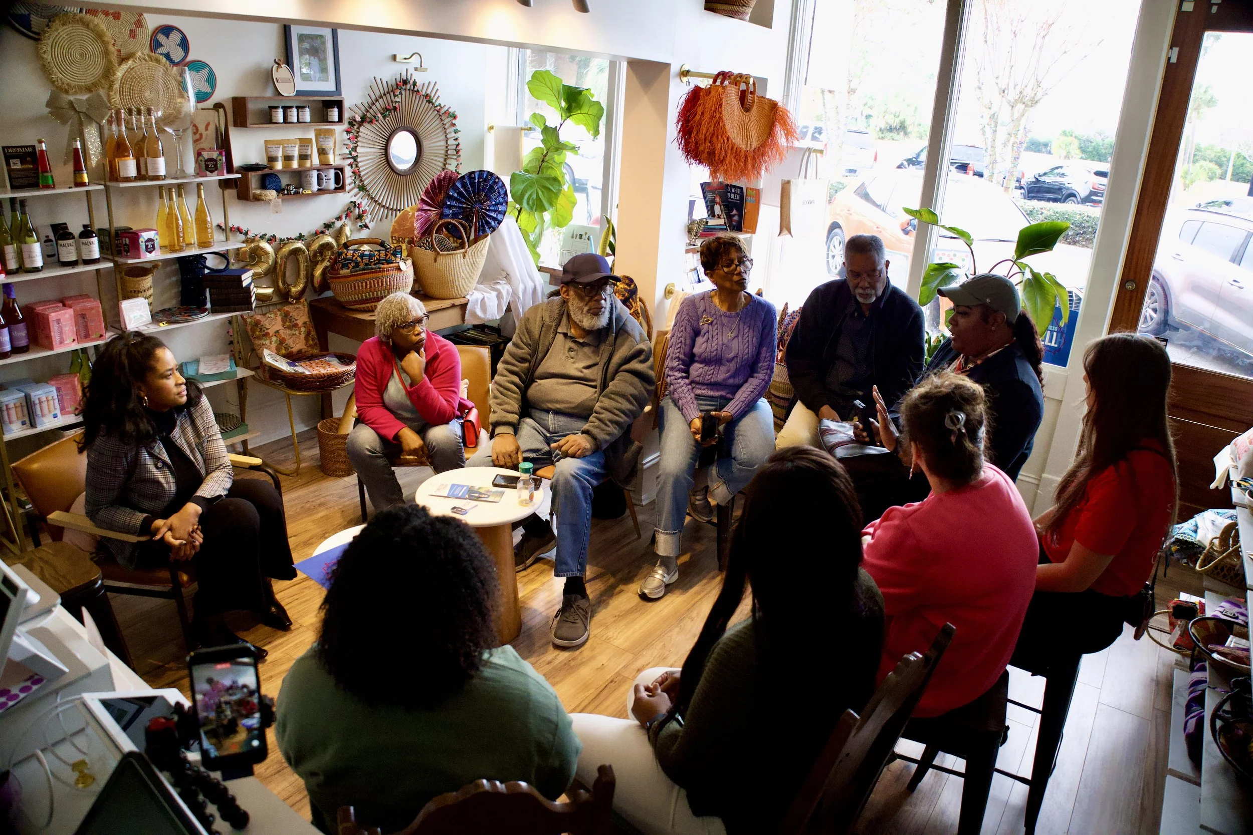 A group of people gathered in a small store or cafe with colorful decorations, shelves of products, and large windows letting in sunlight.