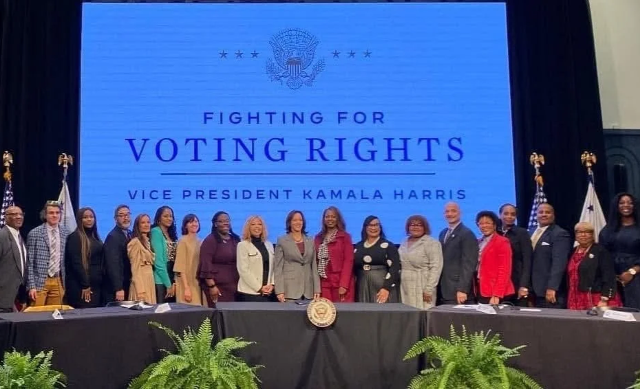 Group of diverse individuals standing on stage in front of a large screen that reads 'Fighting for Voting Rights, Vice President Kamala Harris.' The setting appears to be a formal event or conference.