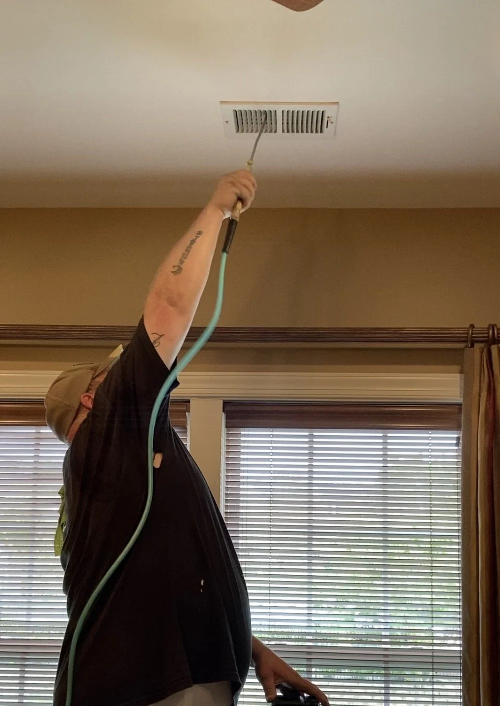 Person cleaning or inspecting an air vent on the ceiling with a tool, standing near windows with blinds, indoors.