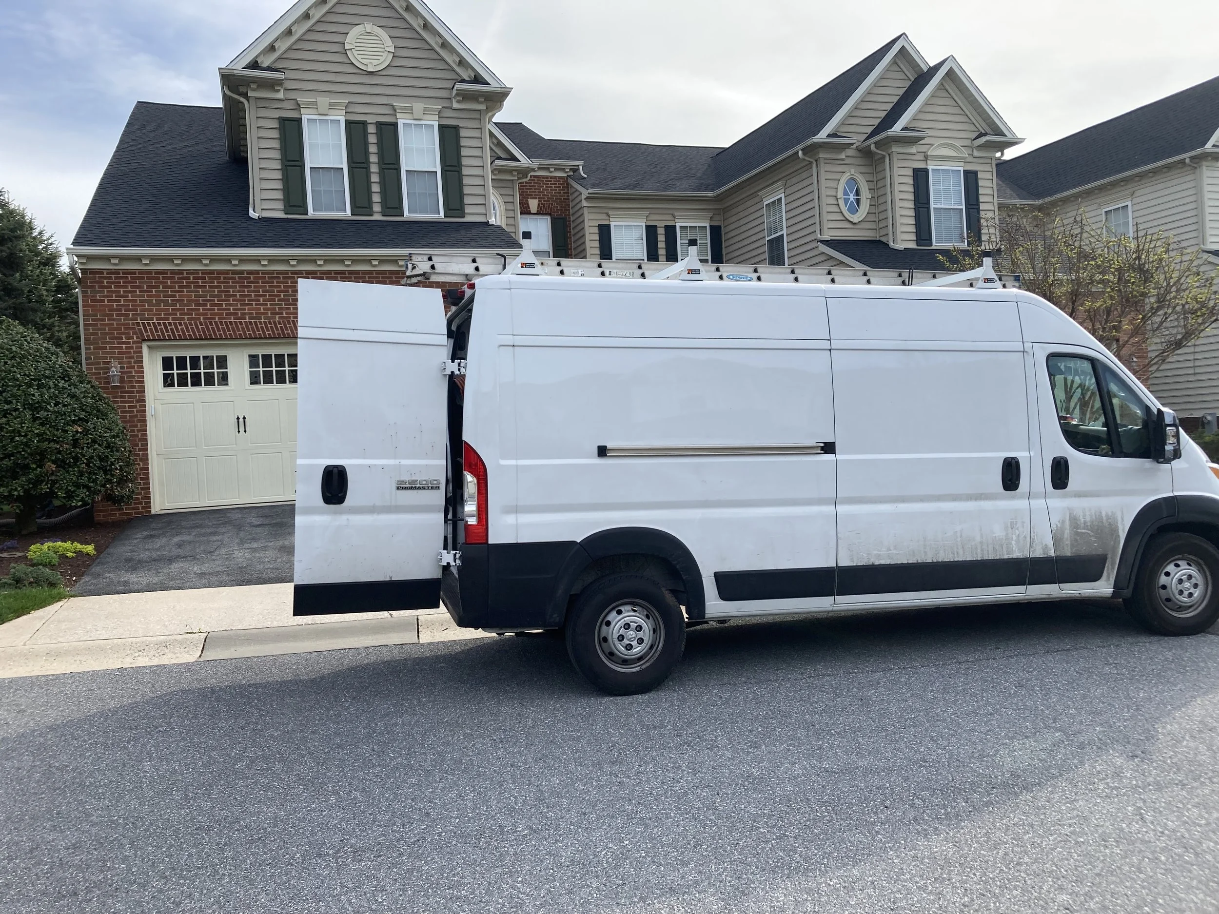A white professional service van parked on the street in front of a suburban house, with a ladder on top of the van.