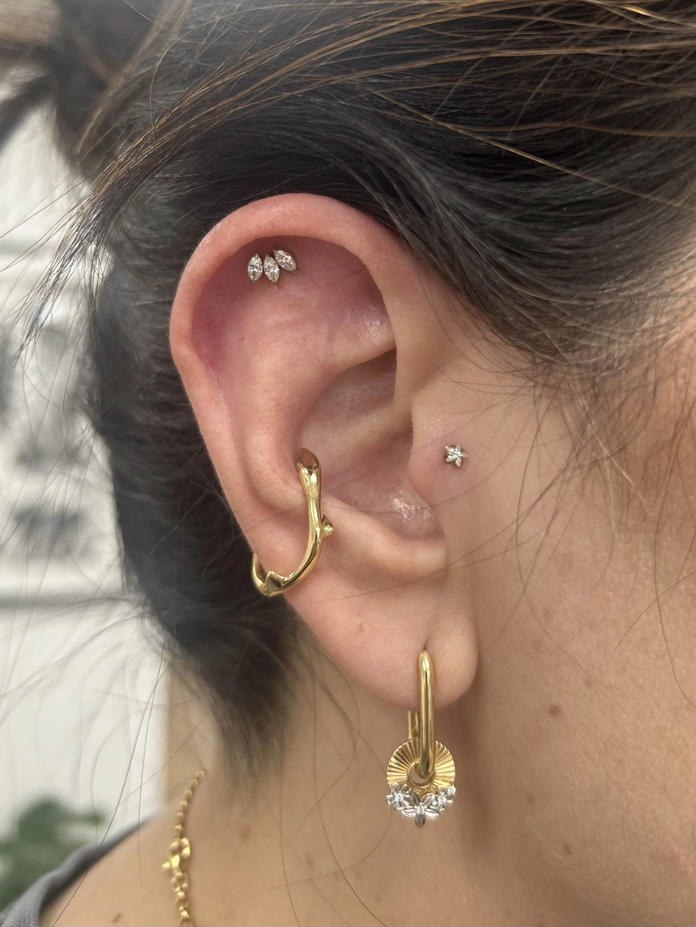 Close-up of a person's ear adorned with multiple earrings, including a small diamond stud, three marquise-shaped diamond earrings, a black and gold hoop, a gold crescent-shaped earring with small diamonds, and a larger gold hoop with black and white details.
