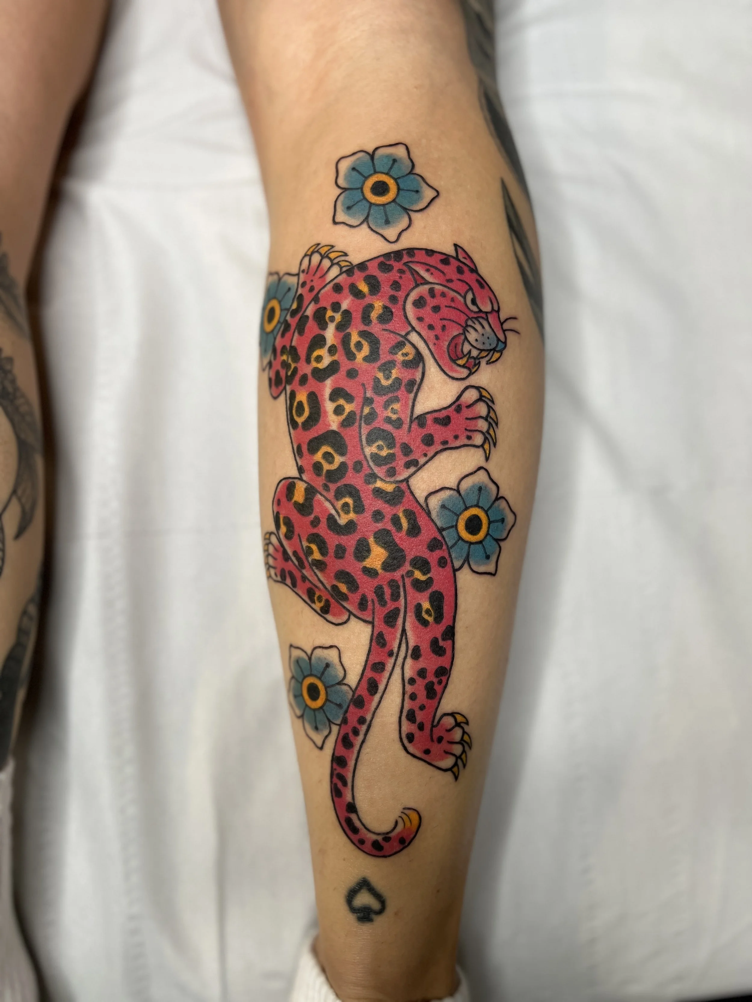 Traditional Tattoo of a pink leopard with black and yellow spots surrounded by blue flowers on a person's arm.