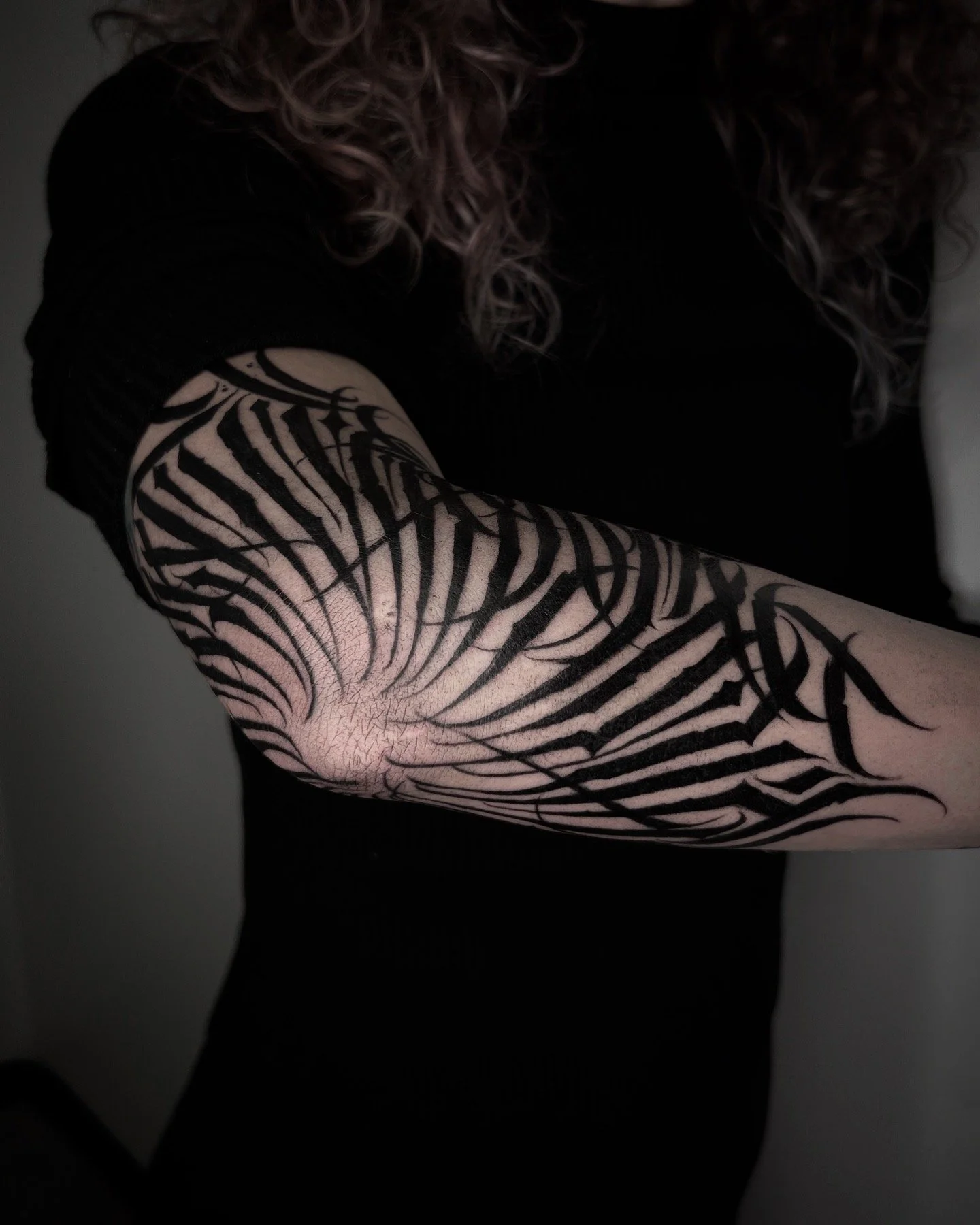 Close-up of a person's arm with a black tattoo sleeve design, featuring lettering, abstract patterns and lines, against a dark background.