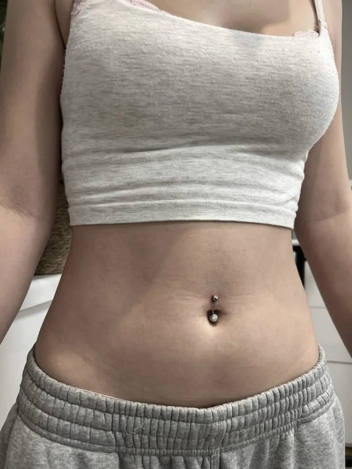 Close-up of a woman's stomach showing a pierced navel with a decorative jewelry, wearing a light gray crop top and matching sweatpants.