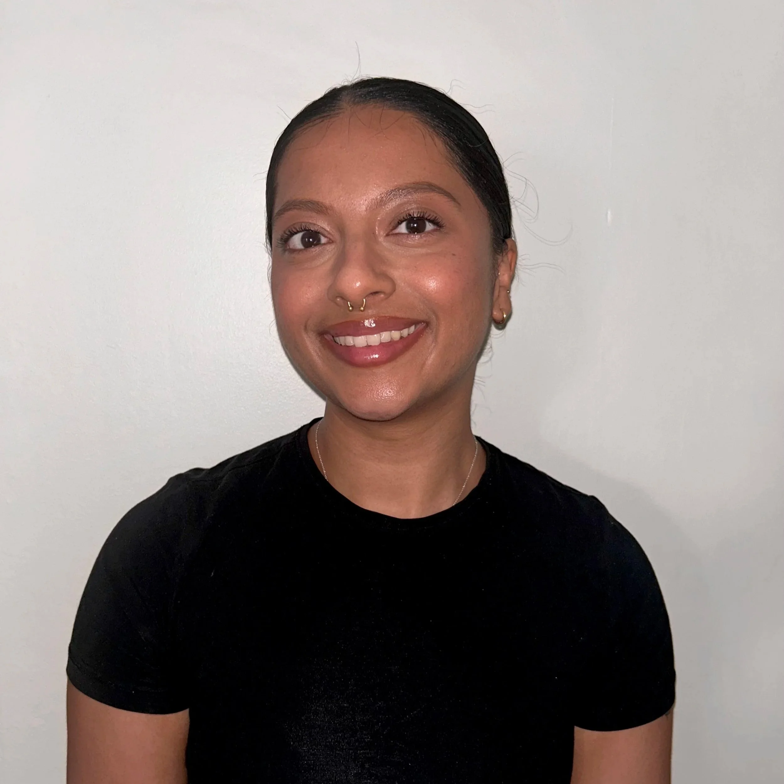 A young woman with dark hair tied back, smiling, wearing a black t-shirt, gold hoop earrings, and a septum piercing, standing against a plain white wall.