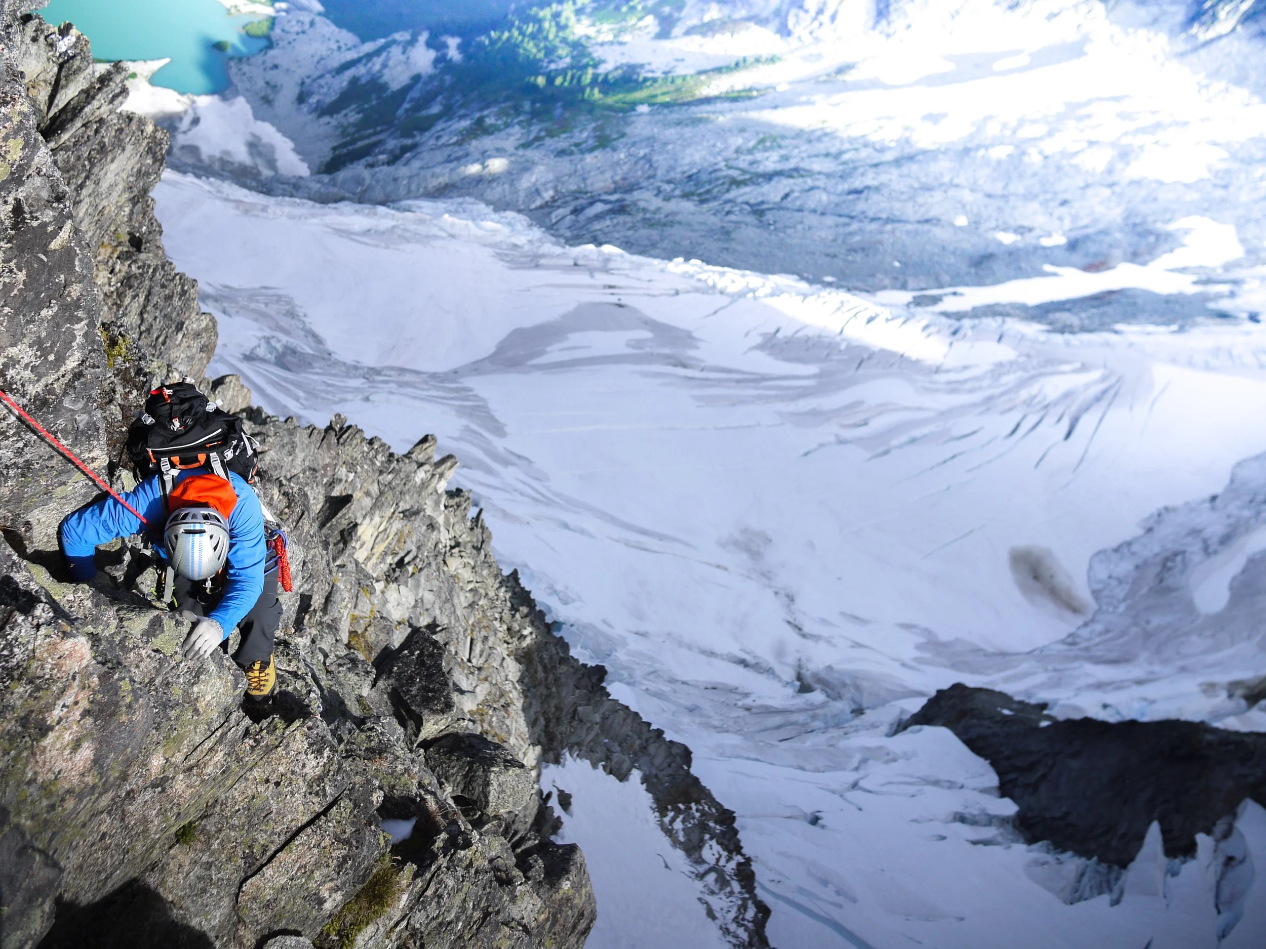 Mountaineer climbing a steep, rocky snow-covered mountain face with icy glacier below.
