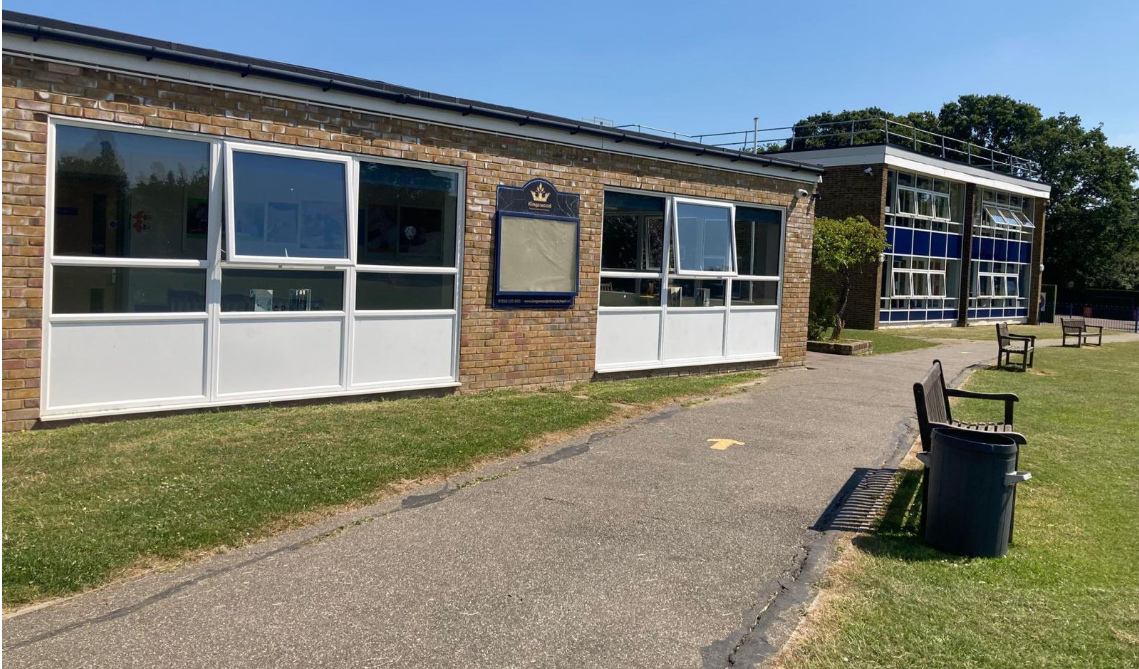 A brick building with a sloped roof, labeled 'LINDON COMMUNITY CENTRE,' situated in a parking lot with grass and bushes, under a clear blue sky.