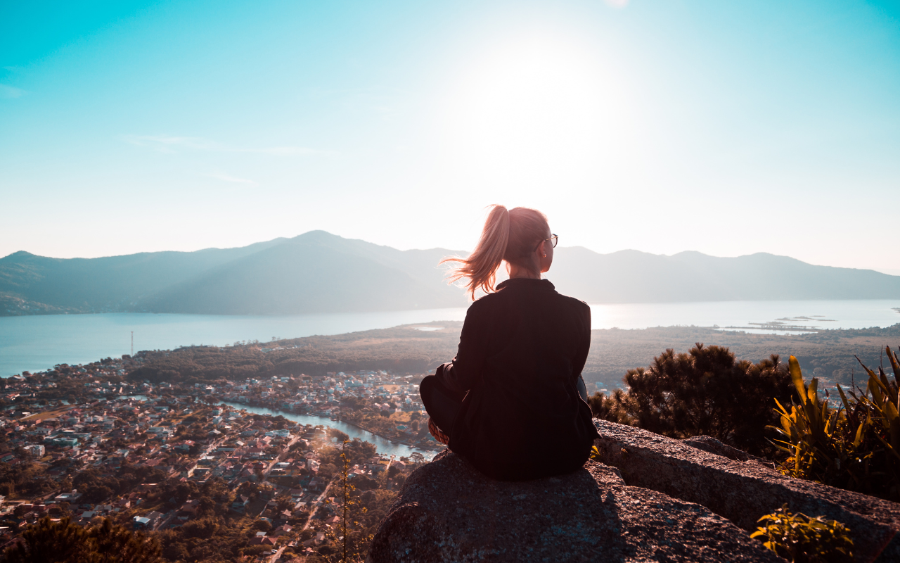 A woman sitting on a large rock overlooking a city, lake, and mountains, with the sun shining in the sky.