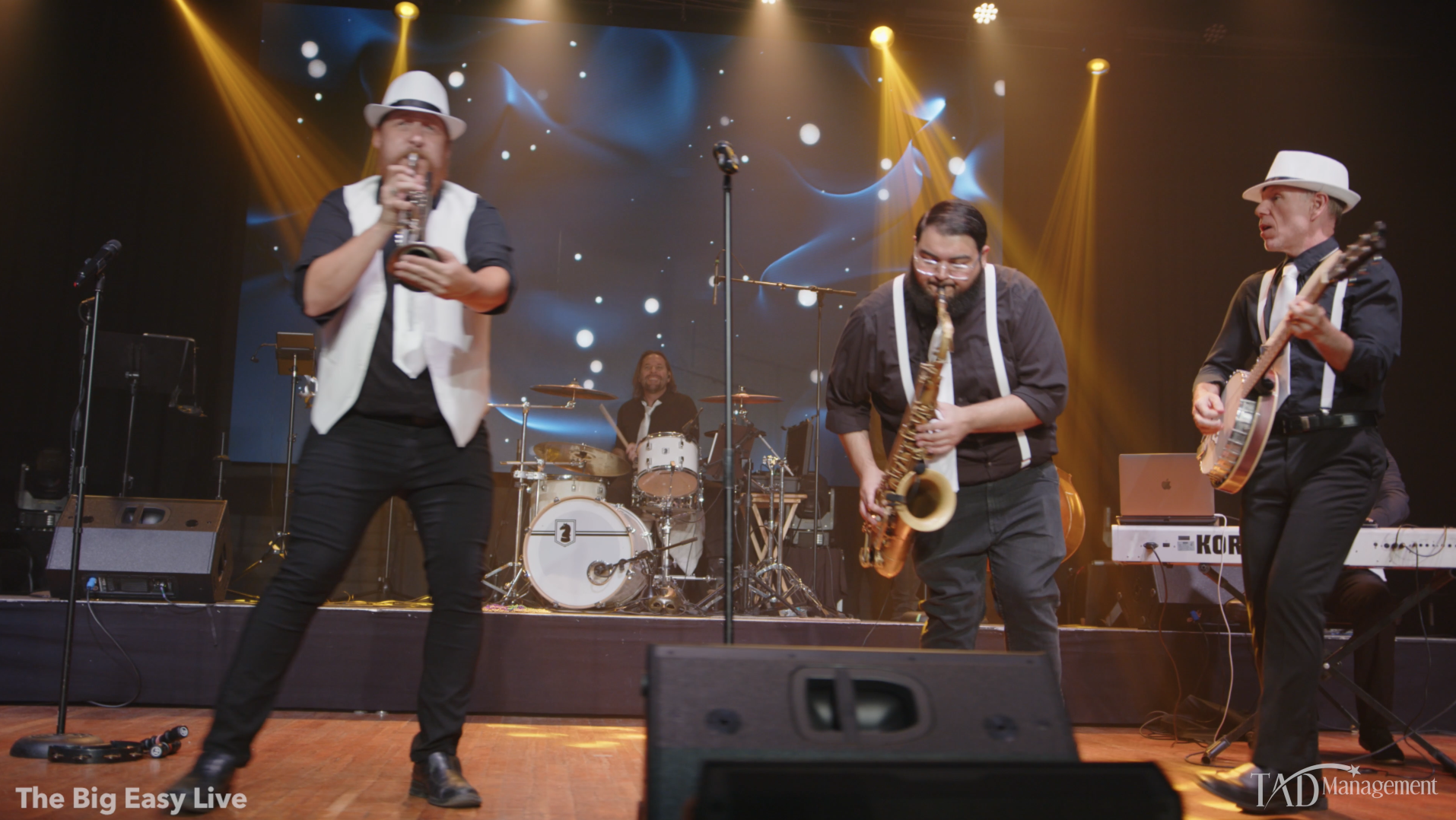 A band performs live on stage with a singer, drummer, saxophonist, guitarist, and keyboardist under stage lights.