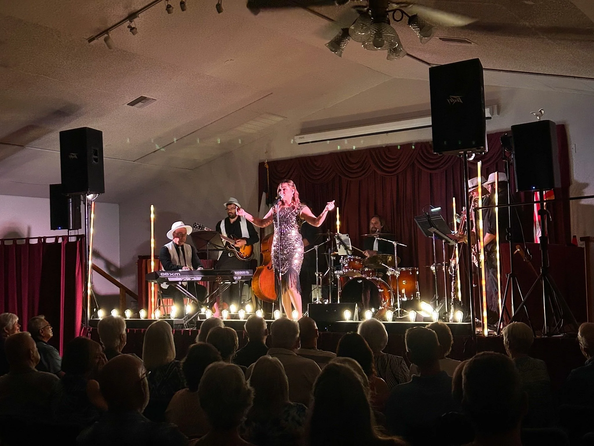 A live band performing on a stage with a singer in a sequined dress, accompanied by a keyboardist, guitarist, drummer, and saxophonist. An audience watches under warm lighting with a red curtain backdrop.