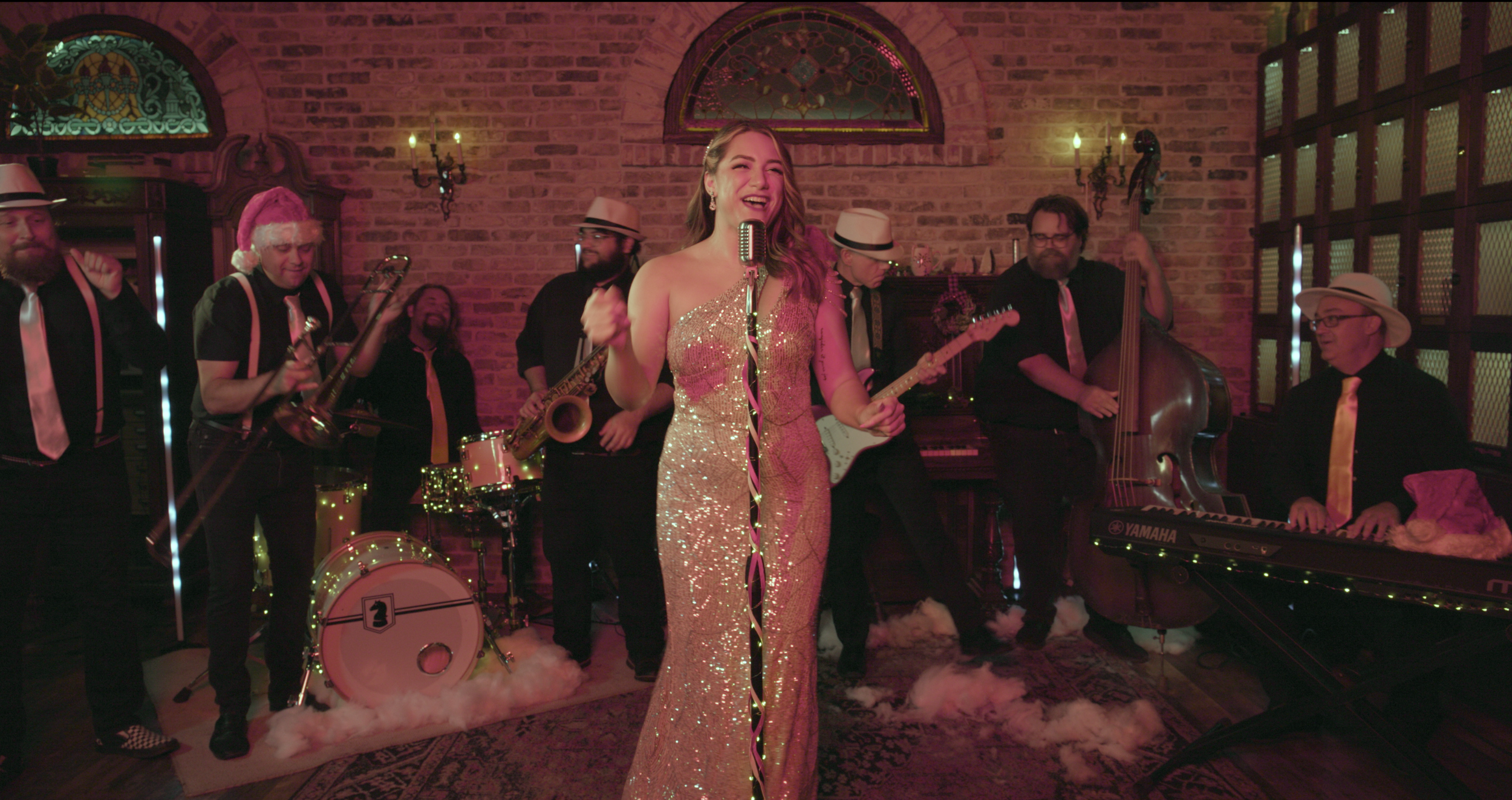 A woman in a sparkling gold dress singing into a microphone surrounded by a jazz band with instruments including drums, saxophone, guitar, double bass, and keyboard, in a warmly lit, brick-walled room with vintage decor.