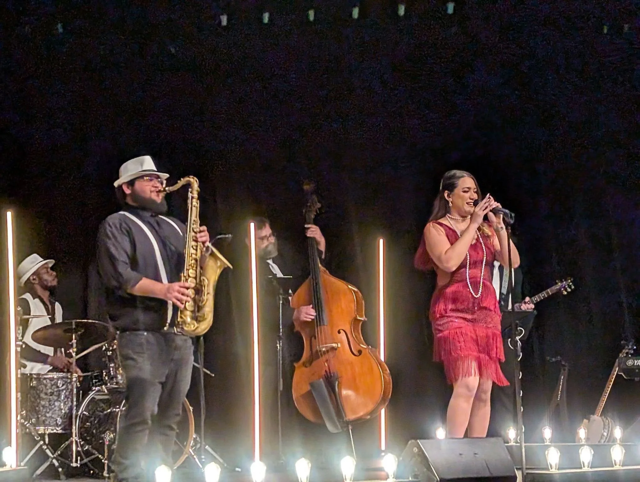 A band performing on stage with a singer holding a microphone, a bassist, a pianist, a drummer, and a guitarist, illuminated by stage lights.