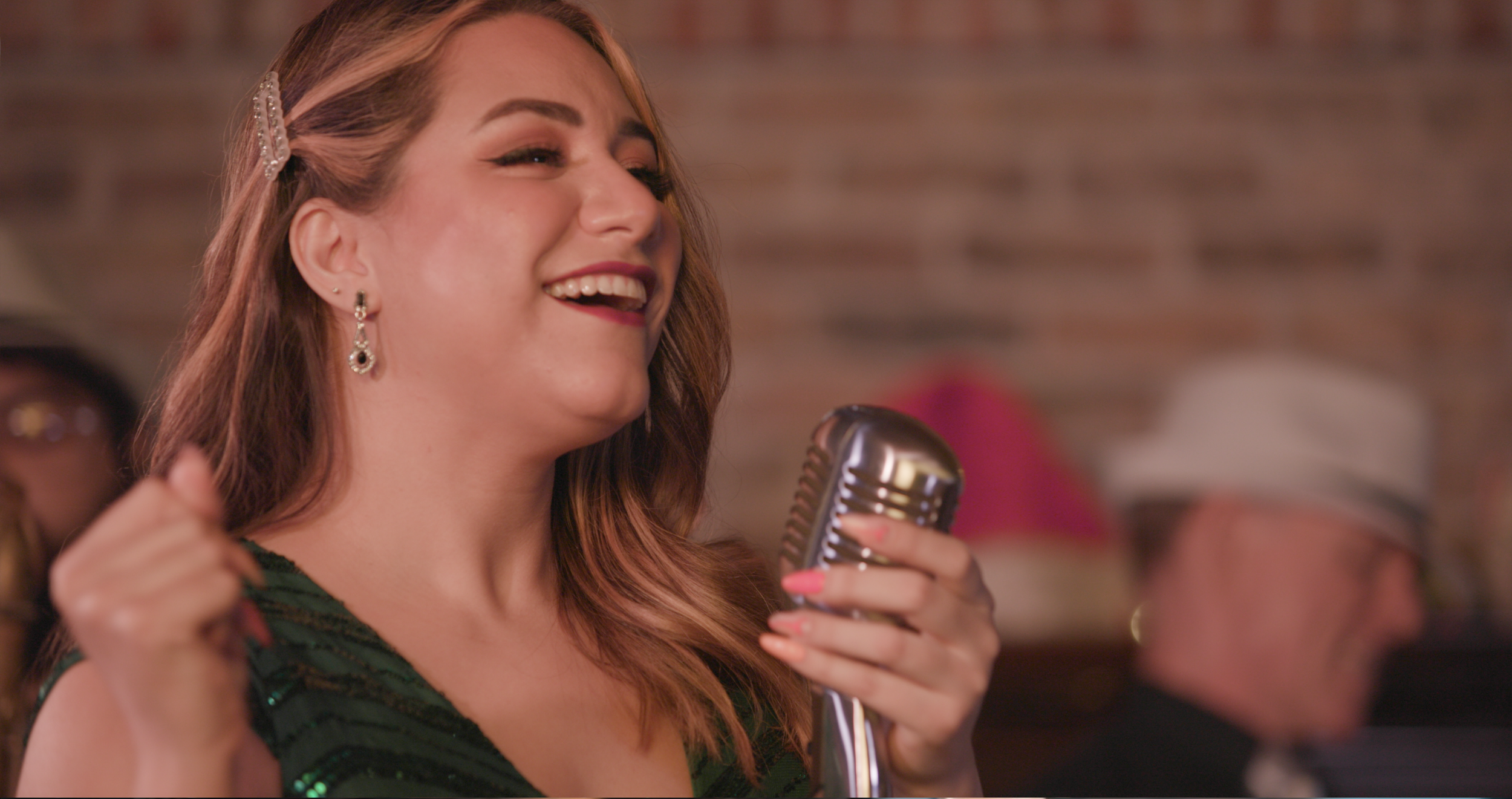 A woman singing into a vintage microphone, smiling with eyes closed, wearing earrings and a green dress. In the background, a man in a tuxedo and white hat listens.