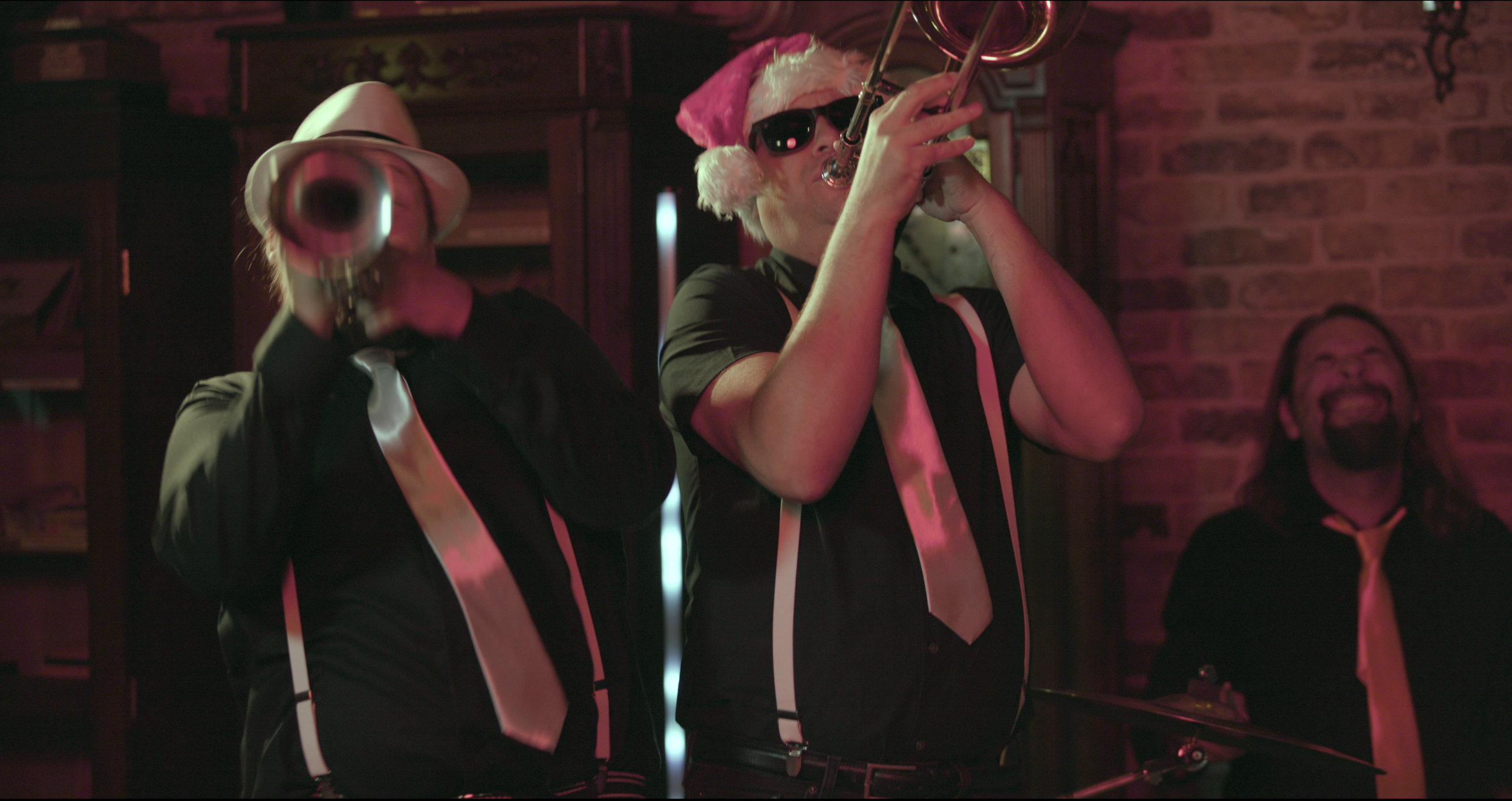 Three men playing musical instruments at a party or celebration, wearing black shirts, white suspenders, and festive accessories like a Santa hat and sunglasses, with a brick wall background.