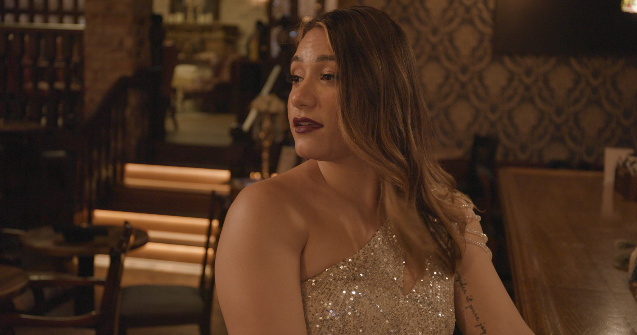 A woman with brown hair wearing a sparkly gold dress sitting in a dimly lit restaurant or bar with patterned wallpaper.