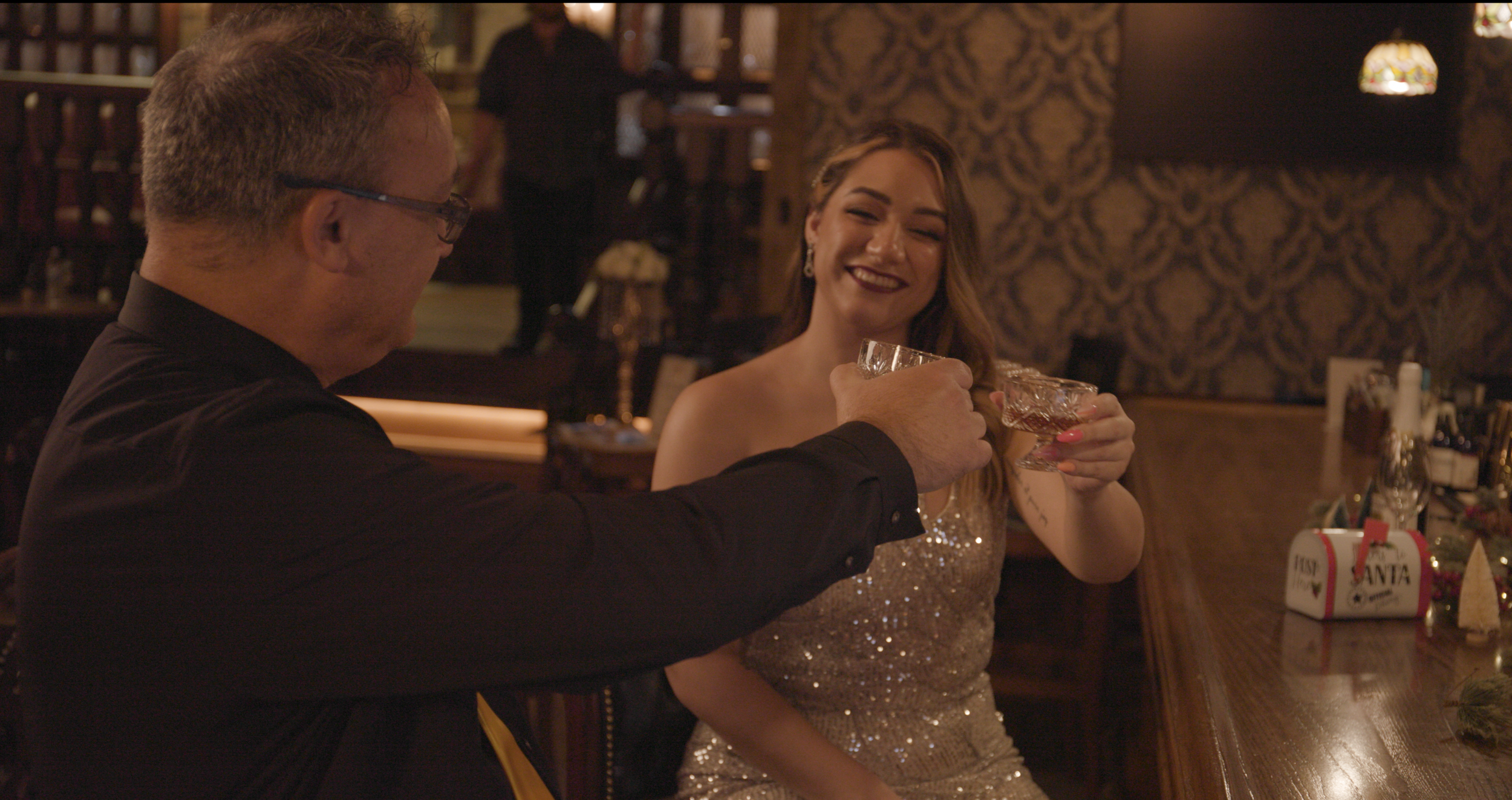 A man and woman clink glasses at a bar, smiling and enjoying drinks in a warmly lit, festive setting with holiday decorations.