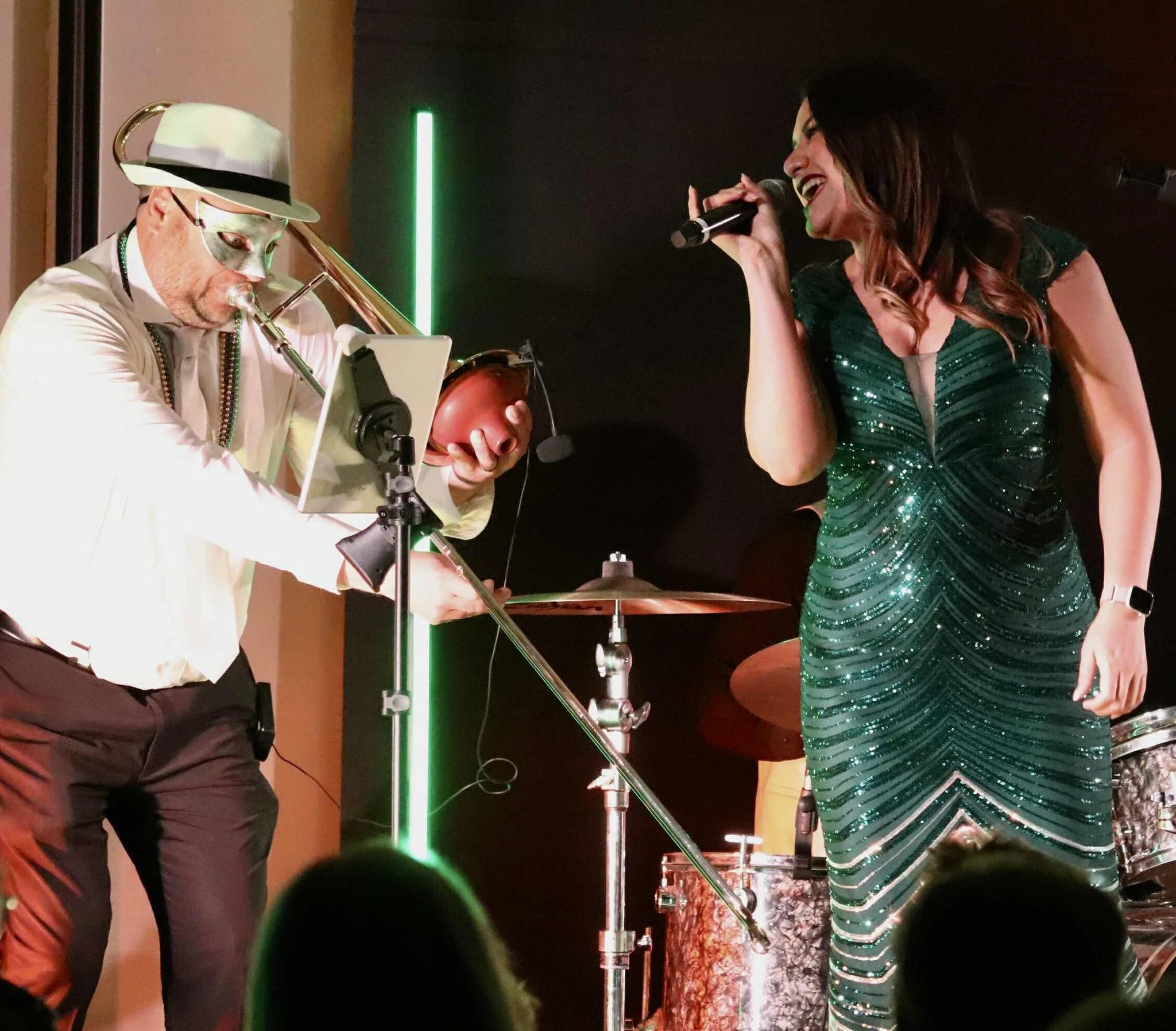 A woman in a green sequined dress singing into a microphone on stage, with a man in a white outfit and silver mask playing the trombone nearby, during a live musical performance.