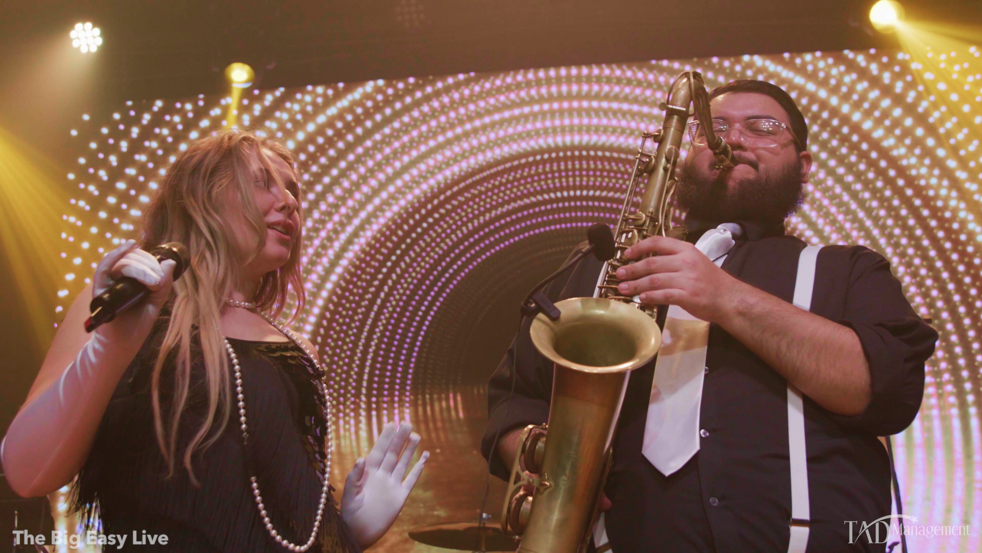 A woman singing into a microphone and a man playing a saxophone on a stage with vibrant, circular, glowing light patterns in the background.