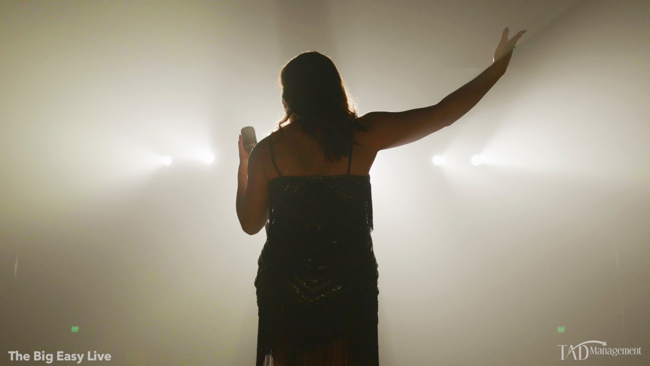 Silhouette of a woman in a black dress holding a microphone on stage, under bright backlights, with one arm raised.