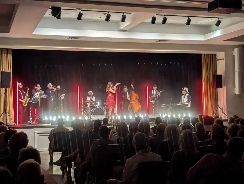 A live band performs on stage with a female singer, surrounded by musicians playing saxophone, drums, double bass, guitar, keyboard, and trumpet, in front of an audience in an indoor venue.