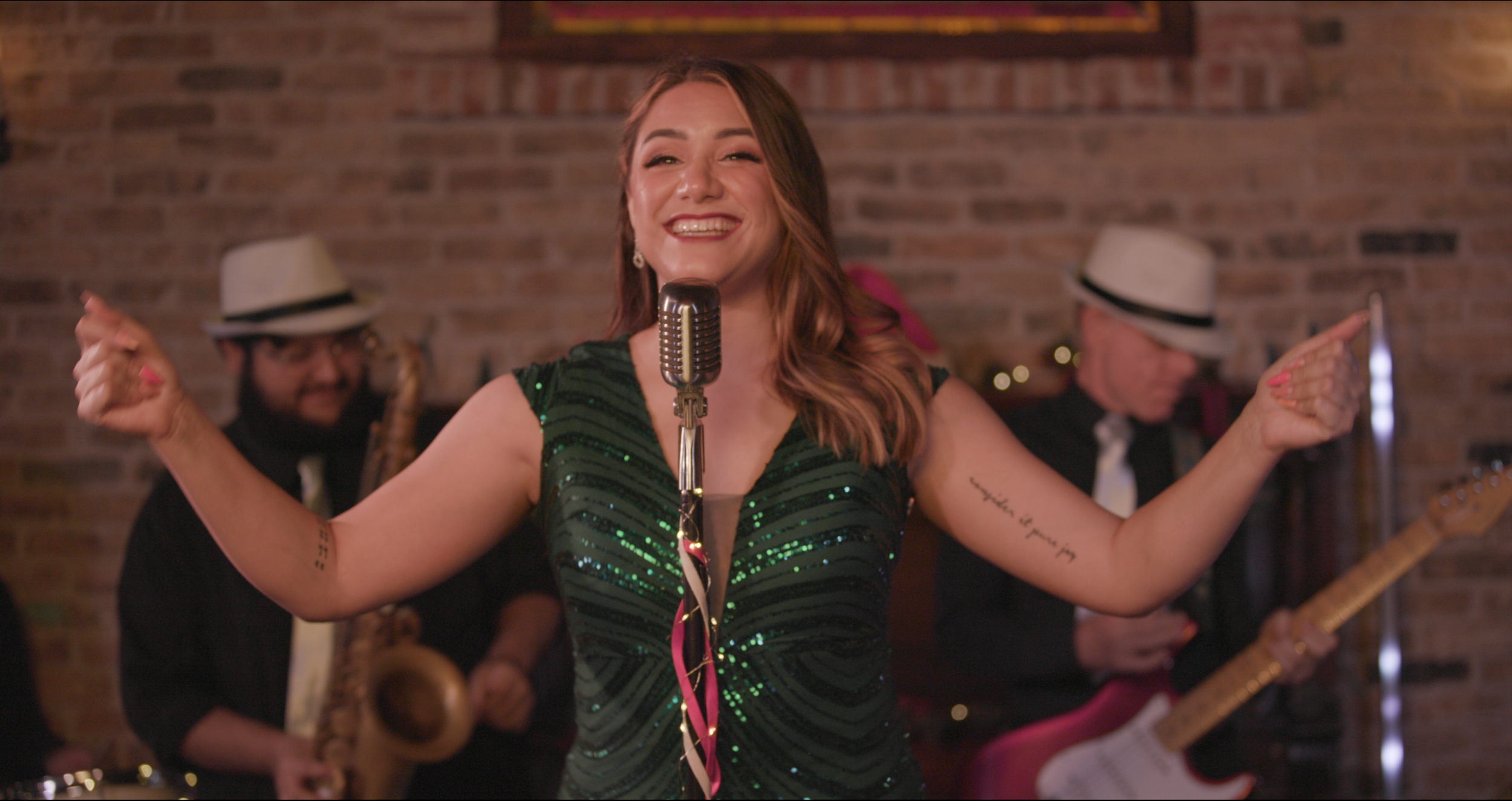 A woman singing into a vintage microphone with a band performing behind her in a cozy brick-walled venue.