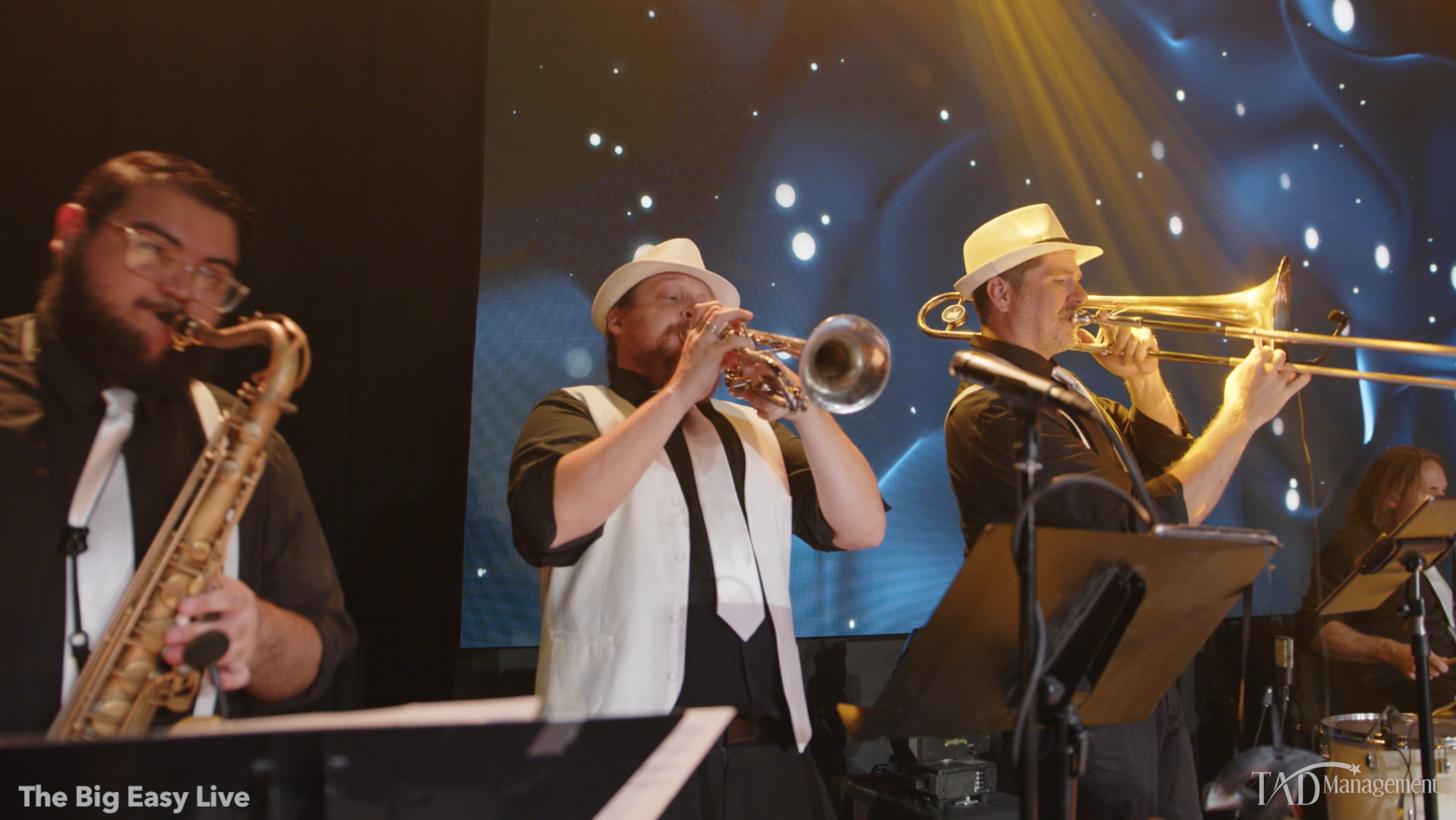 Musicians playing saxophone, trumpet, and trombone on stage during a live performance with a digital backdrop