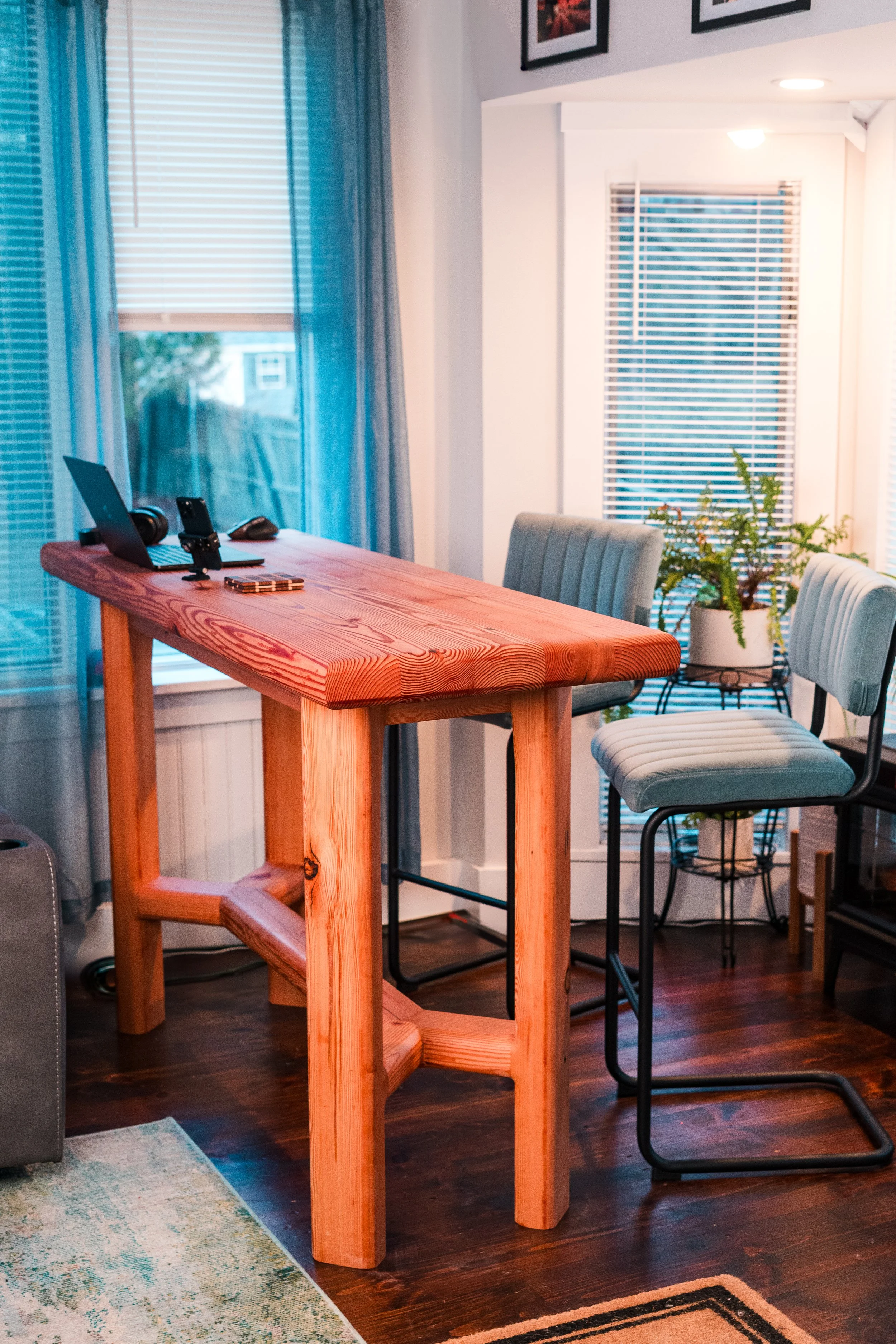 Interior of a cozy dining or workspace area featuring a high wooden table with a natural finish, two light blue cushioned chairs, and potted plants by the window, with framed photos on the wall and natural light coming through the blinds.
