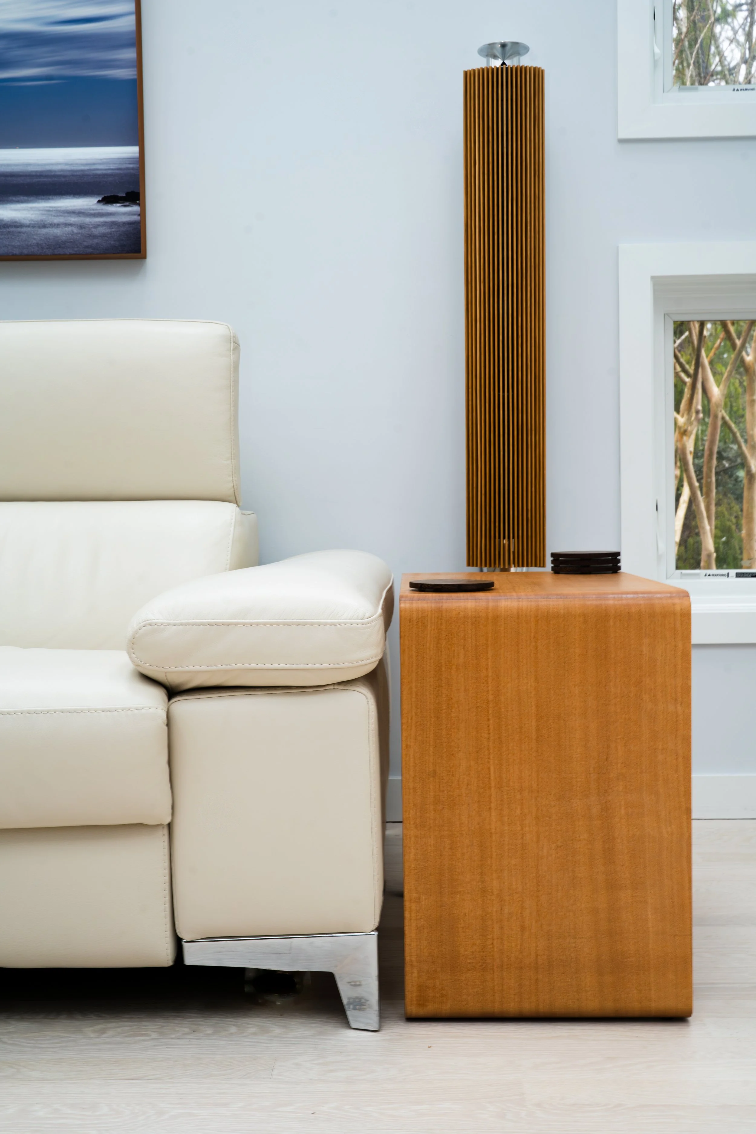 A cream-colored leather sofa placed next to a wooden side table with a tall vertical wooden speaker or decoration on top, and a black item or coaster. Two open windows are visible behind the side table, showing greenery outside. A framed landscape photograph hangs on the white wall to the left.