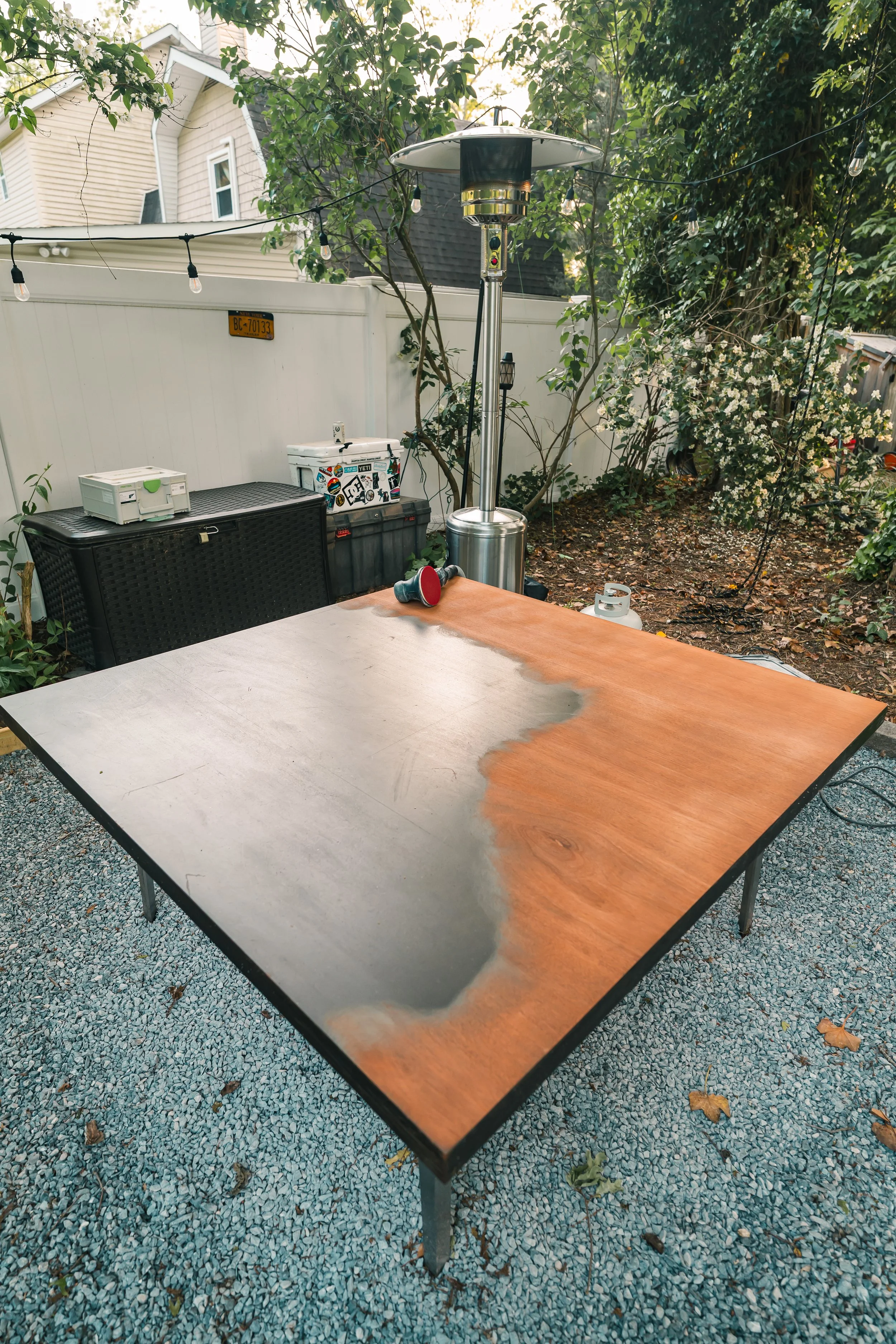 Outdoor patio table with a partially epoxy-resined wood surface, surrounded by trees and outdoor furniture, with string lights overhead.