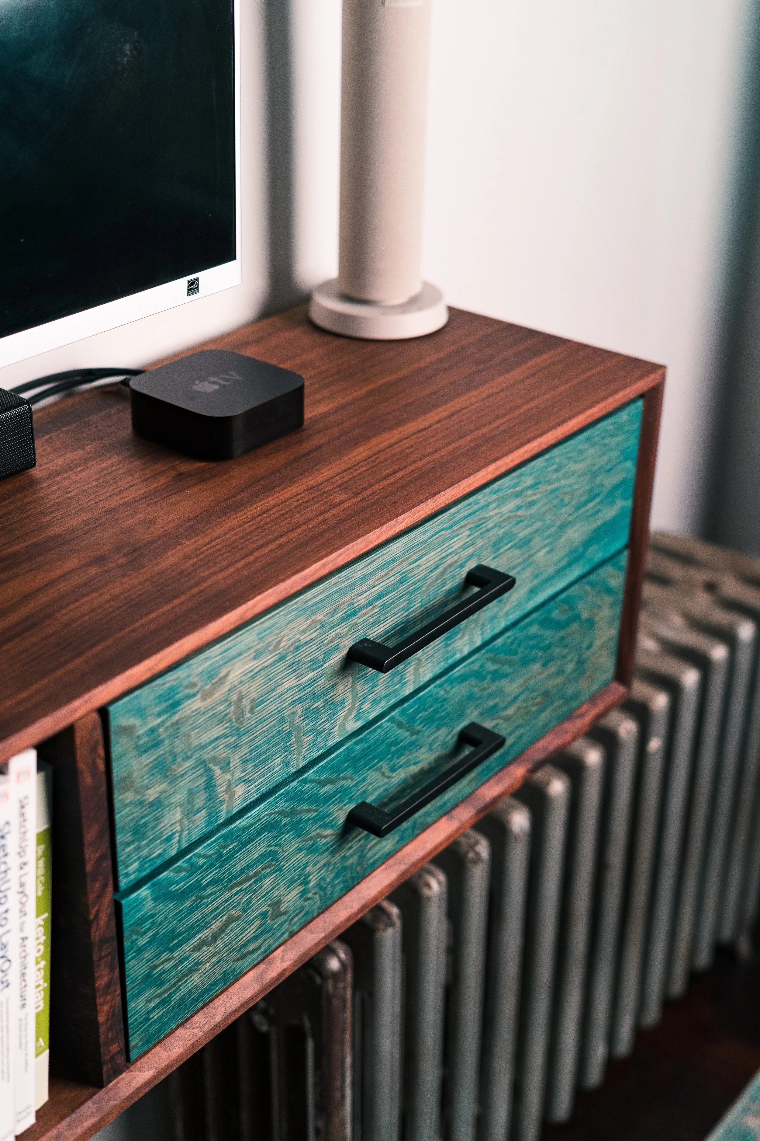 A wooden dresser with a turquoise drawer front and black handles. On top of the dresser, there is a black Apple TV device, a gray speaker, a beige cylindrical lamp, and a few books on the left side. In the background, there is a radiator beneath the dresser.