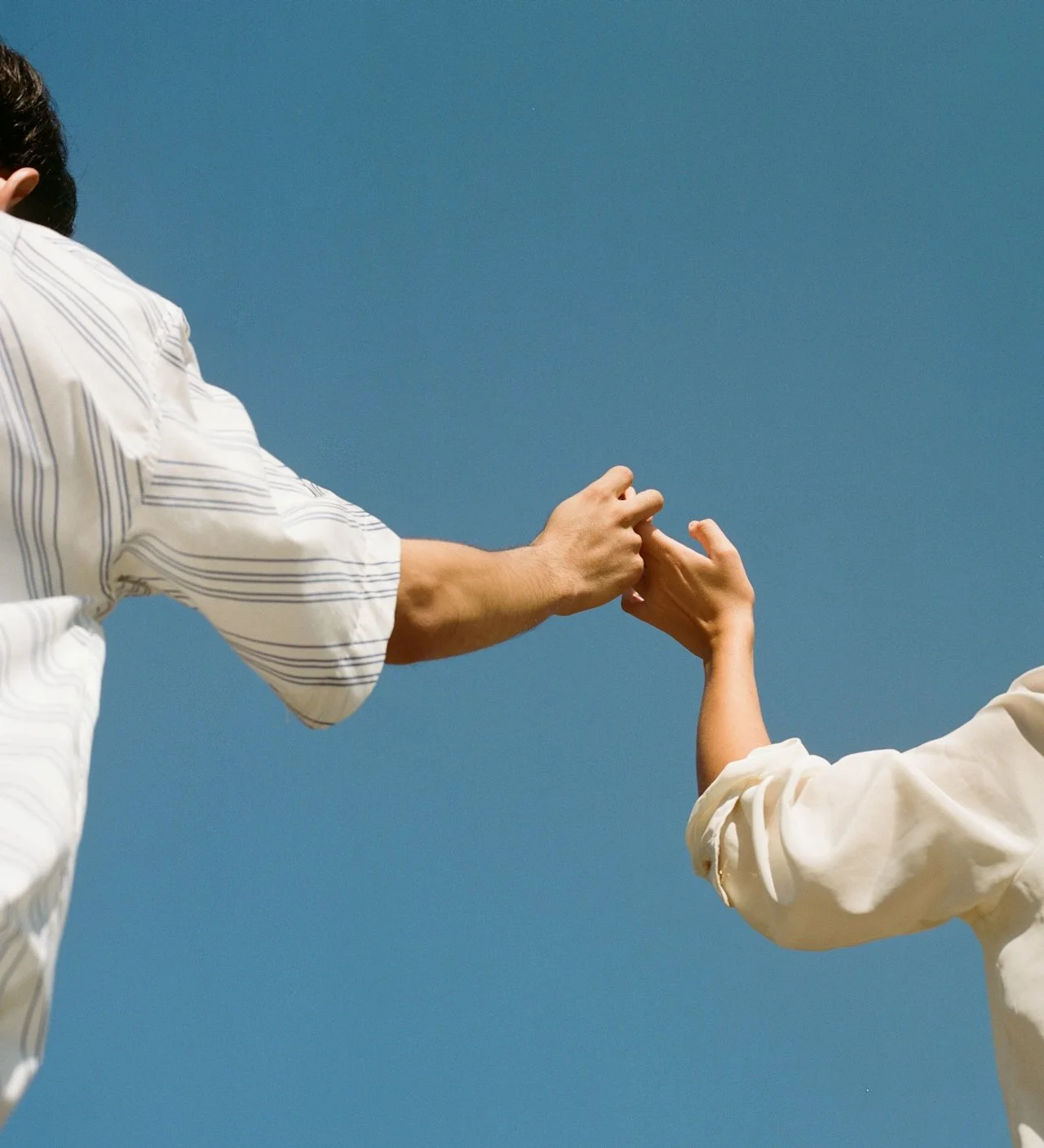 two people reaching toward each other with hands meeting against a clear blue sky