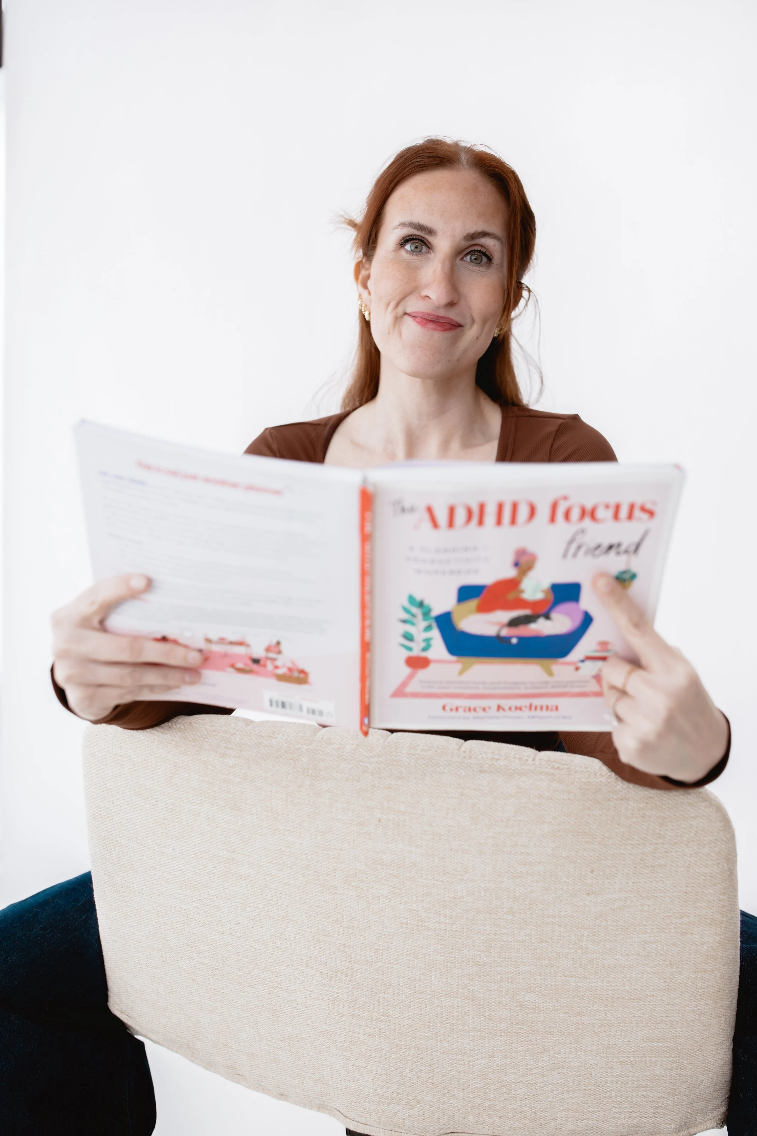 Therapist smiling with an open book, seated casually on a chair, symbolizing welcoming individual therapy for late-diagnosed ADHD adults.