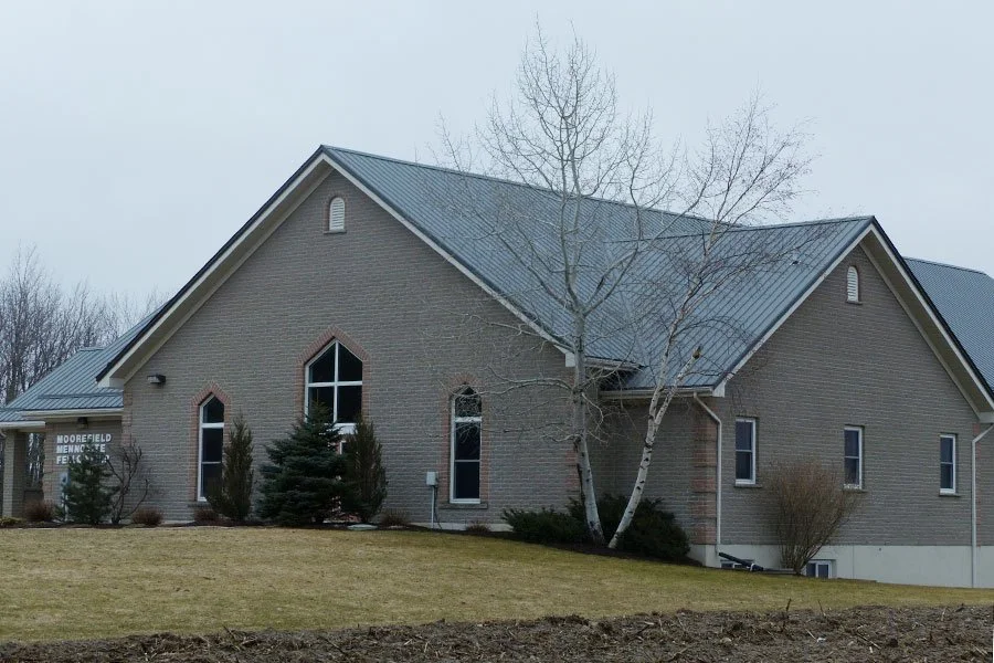 A brick church with a metal roof, leafless trees, and a large grassy front yard.
