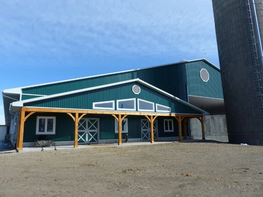 Barn with teal siding, white trim, and a wooden porch supported by beams, located on a dirt lot with a clear blue sky.