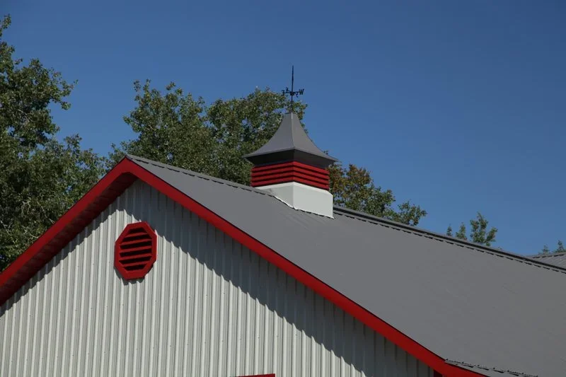 A building with a metallic gray roof, red trim along the edges, a white vent with red slats, and a small cupola with a weather vane, set against a backdrop of trees and blue sky.
