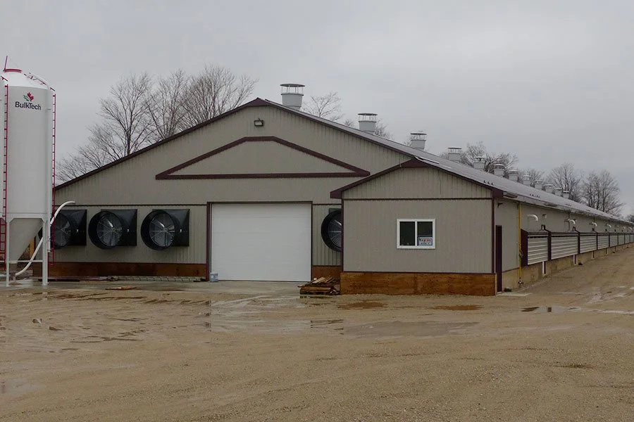A large industrial barn with multiple exhaust fans and a white garage door, on a muddy, overcast day.