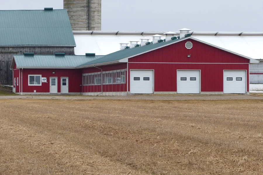 Red barn with three white garage doors and a green roof, located on a farm with dry grass in the foreground.