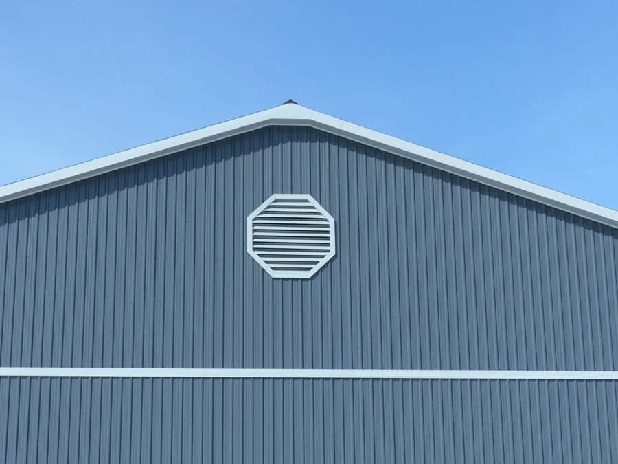Close-up of the upper part of a blue industrial building with vertical metal siding, a white trim, and a roof vent against a clear blue sky.