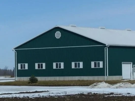 Large green metal barn with a white roof and small windows, located in a rural area with patches of snow on the ground.
