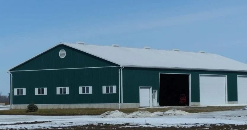 A large green barn with a white roof, partially open garage door, and small windows, set in a rural landscape with patches of snow on the ground and a clear blue sky.
