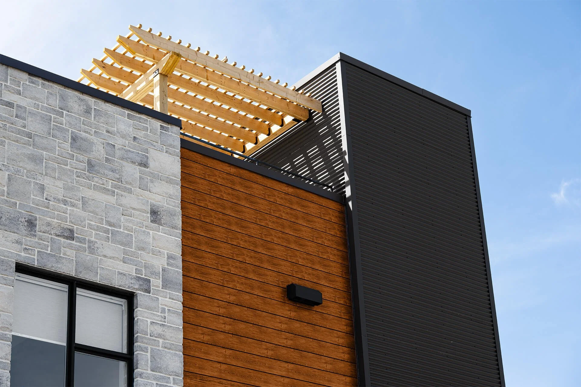 Close-up of a modern building's exterior with gray brick, wooden siding, and black metal paneling, featuring a window and an outdoor wall light, with the roof under construction.