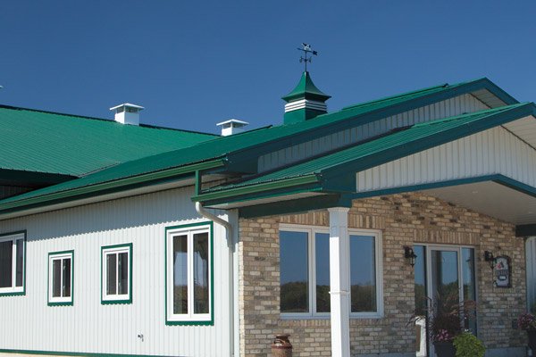 Close-up of a building with a green metal roof, brick and white siding, multiple windows, and a weather vane on top of the roof.