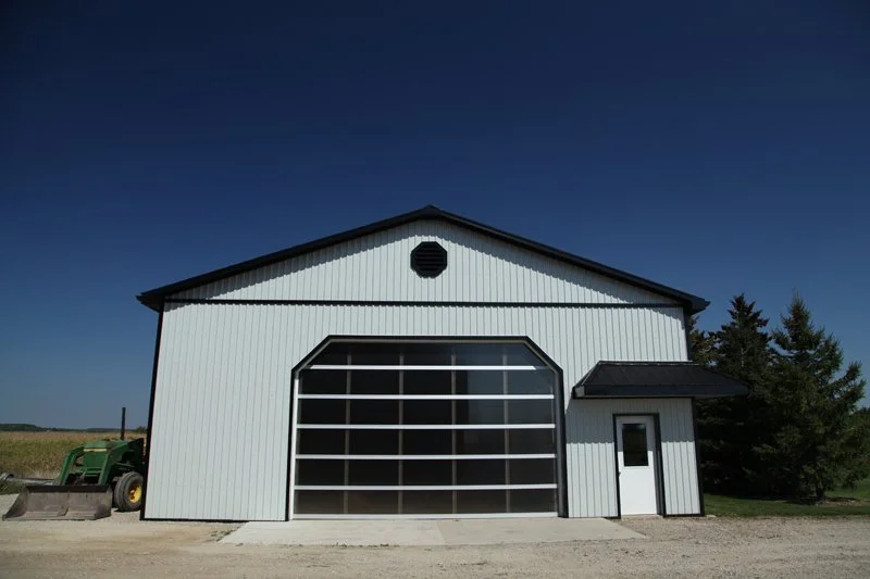 White modern agricultural barn with large glass garage door, small entrance door, and circular vent, set against a clear blue sky and trees.