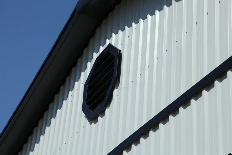 Close-up of a metal building wall with a black octagonal vent and a gutter at the roof edge against a blue sky.